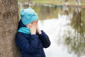 ein einsam traurig Mädchen steht Abdeckung ihr Gesicht mit das Hände von ein Baum auf das Ufer von das See. mental Gesundheit. Teenager Jahre foto