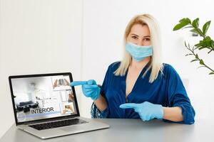 Frau im medizinisch Maske und Gummi Handschuhe sitzt beim Zuhause auf ein Tabelle und funktioniert mit Laptop während Quarantäne. Designer, Künstler, Architekt, Geschäftsmann beim Fernbedienung Arbeit im ein Pandemie Covid. Coronavirus. foto