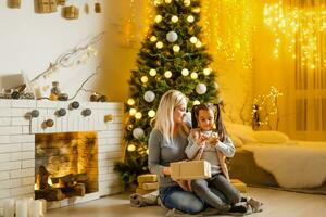 fröhlich Weihnachten und glücklich Ferien heiter Mama und ihr süß Tochter Mädchen austauschen Geschenke. Elternteil und wenig Kind haben Spaß in der Nähe von Weihnachten Baum drinnen. liebend Familie mit die Geschenke im Zimmer. foto