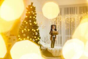 fröhlich Weihnachten und glücklich Ferien heiter süß wenig Kind Mädchen mit gegenwärtig. Kind hält ein Magie Geschenk Box in der Nähe von Weihnachten Baum drinnen. foto