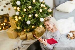 glücklich Frau entspannend beim Weihnachten mit ein Buch wie sie sitzt auf ein Sofa im Vorderseite von das Baum Lachen glücklich beim jemand aus Rahmen foto