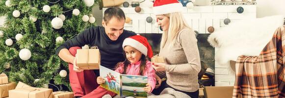 Neu Jahr. Weihnachten. Familie. jung Eltern und ihr wenig Tochter im Santa Hüte sind Ausgaben Zeit zusammen in der Nähe von das Weihnachten Baum beim Zuhause foto