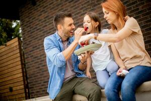 Familie mit ein Mutter, Vater und Tochter Sitzung draußen auf Schritte von ein Vorderseite Veranda von ein Backstein Haus und Essen Erdbeeren foto