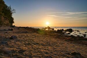 Sonnenuntergang, Stein Strand mit klein und groß Felsen im Vorderseite von das beleuchtet Meer. foto