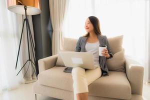 Schönes junges asiatisches Porträt mit Computer und Laptop mit Kaffeetasse auf dem Sofa sitzend foto