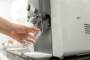 Nahansicht Hand von Frau Füllung Glas mit Wasser von Kühler , Trinken Wasser Maschine foto