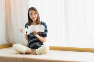 Porträt einer schönen jungen asiatischen Frau, die ein Buch liest und eine Kaffeetasse oder einen Becher auf dem Sofa im Wohnzimmer hält foto