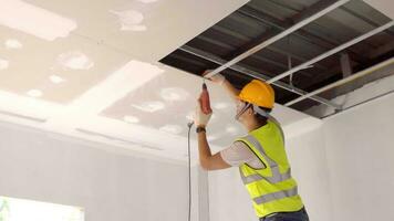 asiatisch Konstruktion Arbeiter im Sicherheit Kleidung und Arbeit Handschuhe ist Befestigung das Trockenbau Decke zu das Metall Rahmen mit ein elektrisch Schraubendreher auf das Decke covere foto