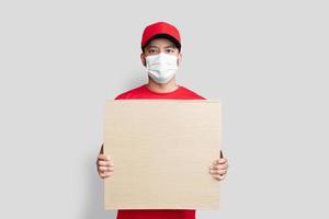 Liefermann-Mitarbeiter in roter Kappe leeres T-Shirt einheitliche Gesichtsmaske halten leeren Karton isoliert auf weißem Hintergrund