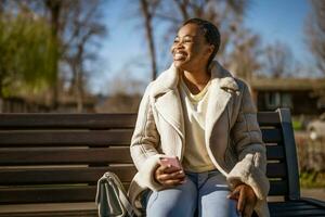 draussen Porträt von glücklich Afroamerikaner Frau auf sonnig Tag. sie ist Sitzung auf Bank im das Straße und mit Smartphone. foto