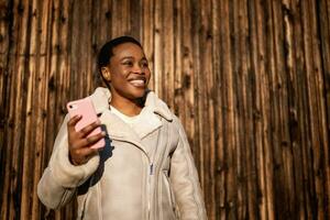draussen Porträt von glücklich Afroamerikaner Frau. sie ist Nachrichtenübermittlung auf Smartphone. foto