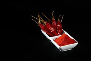 Chilipulver mit rotem Chili in weißer Platte, getrocknete Chilis auf schwarzem Hintergrund