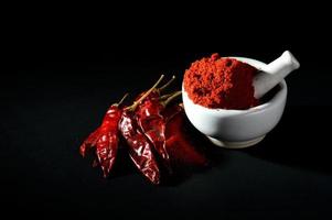 rotes Chilipulver im Stößel mit Mörser und rote Chilischoten auf schwarzem Hintergrund foto