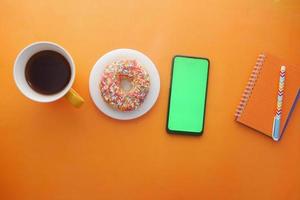 Draufsicht auf Kaffee, Donuts, Notizblock und Smartphone auf orangem Hintergrund foto