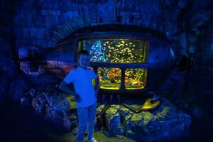 Junge Stand gegen Jahrgang alt unter Wasser U-Boot mit Fische Innerhalb beim Ozeanarium Ausstellungsstück. foto