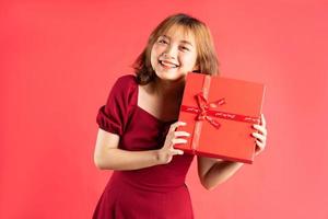 asiatisches junges mädchen im kleid, das rote geschenkbox mit fröhlichem ausdruck im hintergrund hält foto