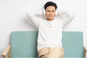 Asiatischer Geschäftsmann, der drahtlose Kopfhörer trägt, um Musik zu hören, während er zu Hause auf dem Sofa sitzt