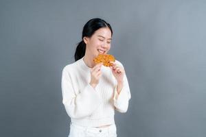 junge asiatische frau trägt einen pullover mit einem glücklichen gesicht und genießt es, gebratenes hähnchen auf grauem hintergrund zu essen foto