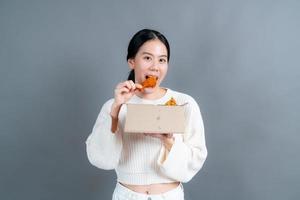 junge asiatische frau trägt einen pullover mit einem glücklichen gesicht und genießt es, gebratenes hähnchen auf grauem hintergrund zu essen foto