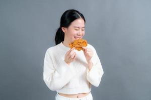 junge asiatische frau trägt einen pullover mit einem glücklichen gesicht und genießt es, gebratenes hähnchen auf grauem hintergrund zu essen foto