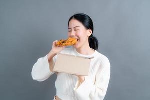 junge asiatische frau trägt einen pullover mit einem glücklichen gesicht und genießt es, gebratenes hähnchen auf grauem hintergrund zu essen foto