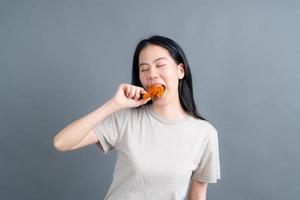 junge asiatische frau trägt einen pullover mit einem glücklichen gesicht und genießt es, gebratenes hähnchen auf grauem hintergrund zu essen foto