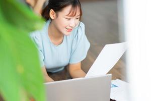 junge asiatische frau, die ein dokument liest und einen laptop benutzt, wenn sie von zu hause aus arbeitet