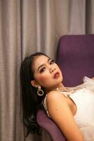 ein asiatisch Frau im ein sexy Weiß Kleid ist Lügen auf ein Sofa mit glatt Beine im ein Hotel Zimmer foto