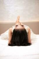 ein asiatisch Frau im ein Weiß Kleid ist Lügen auf ein Weiß Bett mit ihr schwarz Haar lose im ein Hotel foto
