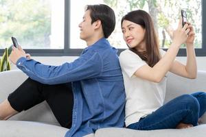 Junges asiatisches Paar sitzt mit dem Rücken aufeinander und benutzt das Telefon auf dem Sofa, das Mädchen schaut auf das Telefon ihres Freundes foto