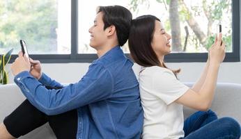 Junges asiatisches Paar sitzt mit dem Rücken aufeinander und benutzt das Telefon auf dem Sofa