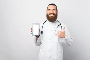 glücklicher junger Arzt mit Bart, der einen leeren Bildschirm des Tablets zeigt und den Daumen über der weißen Wand zeigt foto