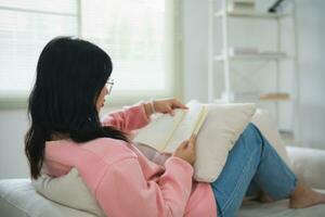 froh glücklich asiatisch Frau lächelnd und lesen Buch während Sitzung auf Couch Sofa im Leben Zimmer beim heim. asiatisch Frauen entspannen lesen Buch Lächeln beim Sofa im das Haus. Aktivität Hobby beim Haus Konzept. foto