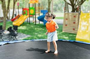 asiatisch Kind Mädchen ist Springen auf Trampolin auf Spielplatz Hintergrund. glücklich Lachen Kind draußen im das Hof auf Sommer- Urlaub. springen hoch auf Trampolin. Aktivität Kinder im das Kindergarten Schule. foto