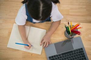 Draufsicht. asiatisch Baby Mädchen lächelnd verwenden Laptop oder Schreiben Zeichnung Farbe auf Hinweis Buch Studie online auf Holz Tabelle Schreibtisch im Leben Zimmer beim heim. Bildung Lernen online von Zuhause Konzept. foto