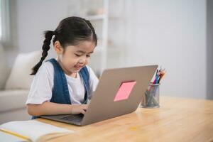 asiatisch Baby Mädchen lächelnd verwenden Laptop oder Schreiben Zeichnung Farbe auf Hinweis Buch Studie online auf Holz Tabelle Schreibtisch im Leben Zimmer beim heim. Bildung Lernen online von Zuhause Konzept. foto