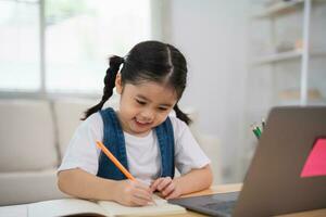 asiatisch Baby Mädchen lächelnd verwenden Laptop oder Schreiben Zeichnung Farbe auf Hinweis Buch Studie online auf Holz Tabelle Schreibtisch im Leben Zimmer beim heim. Bildung Lernen online von Zuhause Konzept. foto