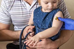 Impfung eines kleinen Mädchens in den Armen ihres Vaters in der Arztpraxis gegen das Coronavirus. lustiges klebepflaster für kinder. Impfung gegen Covid-19, Grippe, Infektionskrankheiten.