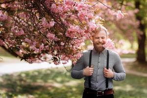 bärtiger Mann mit stilvollem Haarschnitt mit Sakura-Blumen im Hintergrund. Hipster in Hosenträgern in der Nähe mit Zweig von Sakura-Blüten in den Zähnen. Harmonie mit dem Naturkonzept. foto