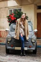 schönes Mädchen in der Stadt. glückliche junge Frau, die in der Nähe eines Retro-Autos mit Weihnachtsbaum steht. kalter glücklicher Wintertag. Urlaub, Weihnachten, Winter, Liebe, Beauty-Konzept foto