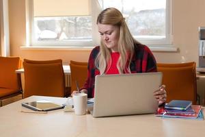 lächelnde blonde Frau sitzt an einem Laptop, trinkt Tee und arbeitet. foto