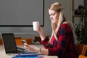 lächelnde blonde Frau sitzt an einem Laptop, trinkt Tee und arbeitet. foto