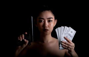 asiatische Frau mit einer Hand, die eine Waffe und Geld auf dem schwarzen Hintergrund hält