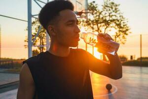 schwarz Mann tun Sport im Morgen, Trinken Wasser auf Basketball Gericht auf Sonnenaufgang foto