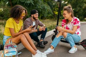 glücklich jung Unternehmen von lächelnd freunde Sitzung im Park auf Gras mit elektrisch trete Roller foto