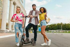 glücklich jung Unternehmen von lächelnd freunde Gehen im Straße mit elektrisch trete Roller foto