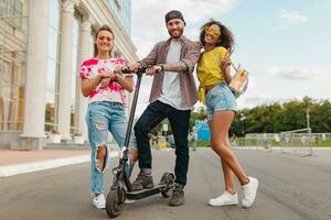 glücklich jung Unternehmen von lächelnd freunde Gehen im Straße mit elektrisch trete Roller foto