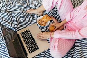 junge hübsche blonde frau in rosa pyjamas sitzt auf dem bett und arbeitet am laptop, freiberufler zu hause foto