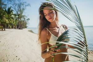 jung dünn Frau im Weiß Bikini Badebekleidung halten Blatt von Palme Baum foto