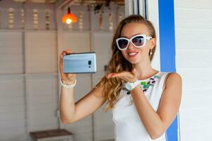 attraktiv Frau im Weiß Kleid im Sommer- Cafe im Sonnenbrille mit Telefon nehmen Foto